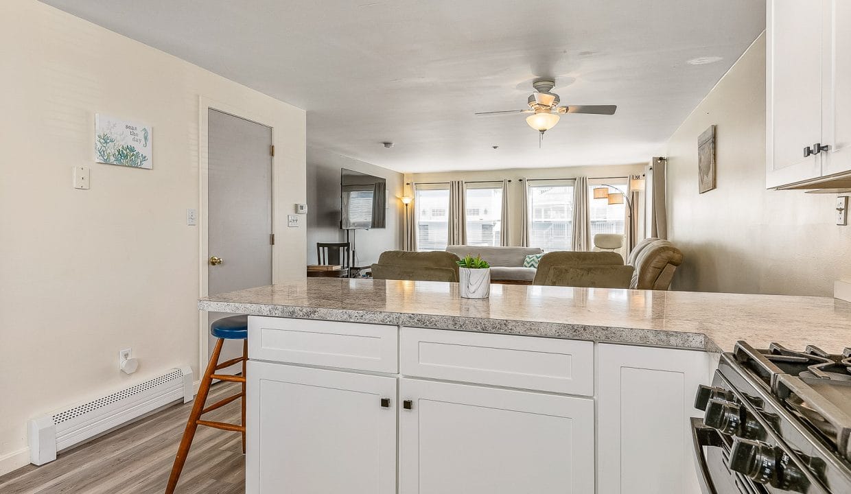 Modern open-concept kitchen and living room with white cabinetry, granite countertops, a ceiling fan, and natural light from large windows.