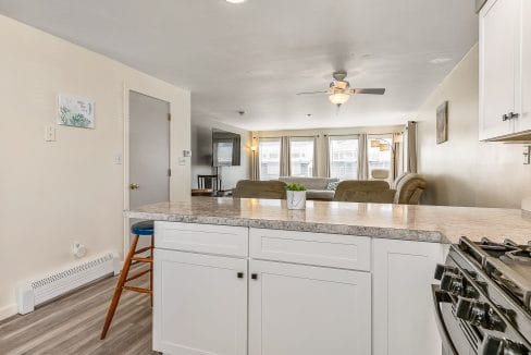 Modern open-concept kitchen and living room with white cabinetry, granite countertops, a ceiling fan, and natural light from large windows.