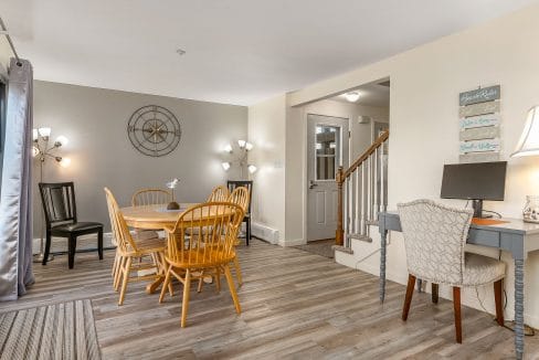 Dining room with a round wooden table and chairs, adjacent to a small work desk with a computer. Stairs lead to another level. Decorative wall elements and lamps are visible.