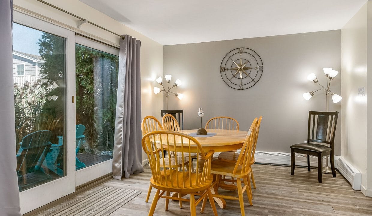 Dining area with a round wooden table, six chairs, and a large decorative clock on the wall. Two modern light fixtures are present, and a sliding door opens to an outdoor space with chairs.