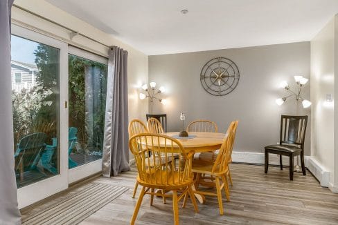 Dining area with a round wooden table, six chairs, and a large decorative clock on the wall. Two modern light fixtures are present, and a sliding door opens to an outdoor space with chairs.