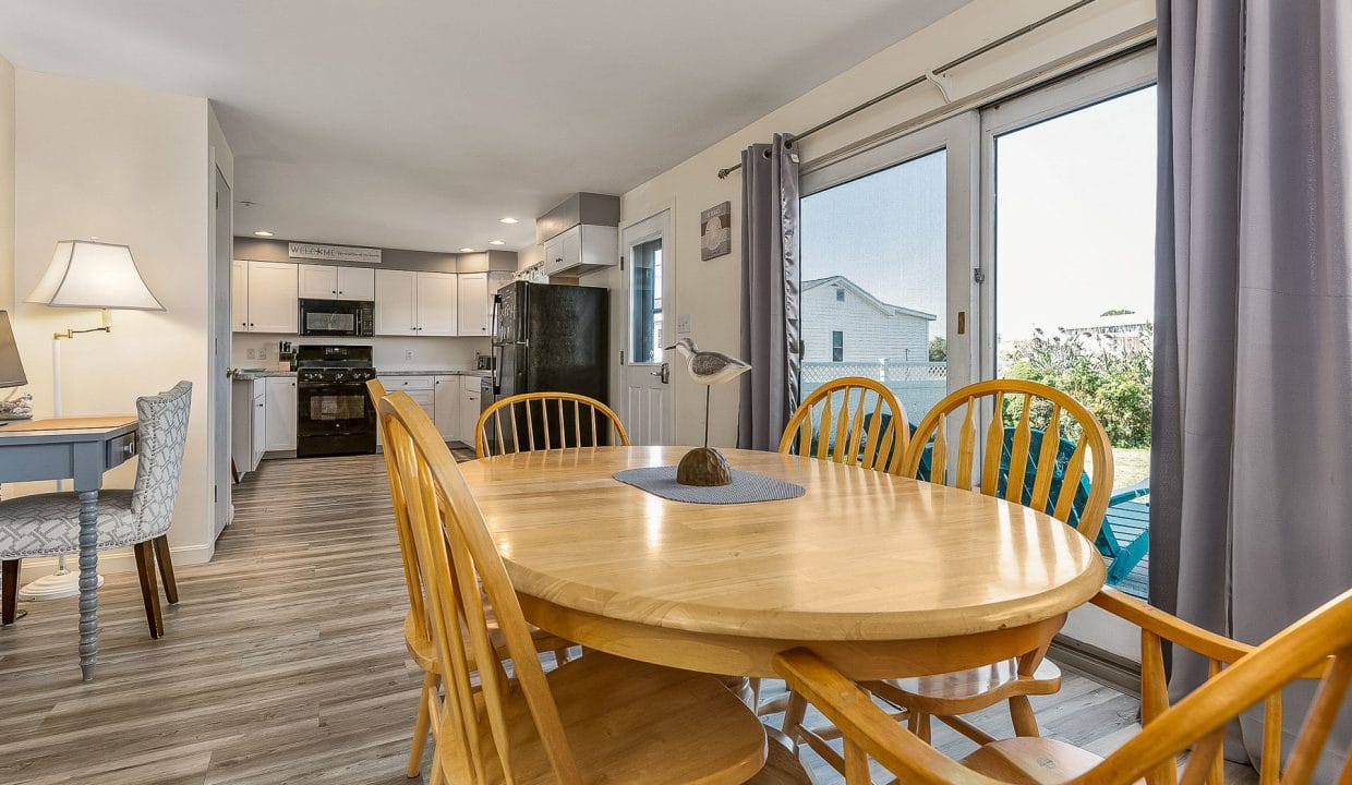 Dining area with a wooden table and chairs, near large windows. Kitchen with white cabinets and a refrigerator visible in the background. Floor is light wood.