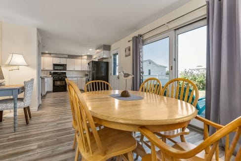 Dining area with a wooden table and chairs, near large windows. Kitchen with white cabinets and a refrigerator visible in the background. Floor is light wood.