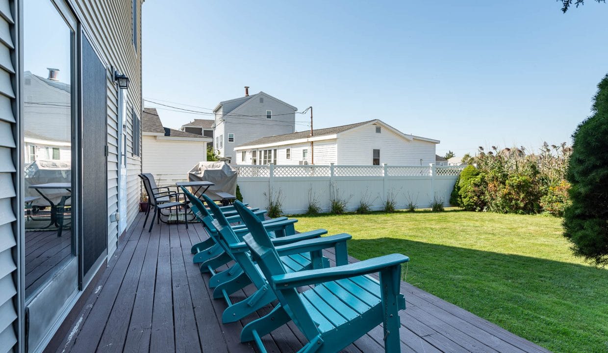 Backyard with wooden deck, teal Adirondack chairs, and a grassy lawn. White houses and a fence are visible in the background under a clear blue sky.