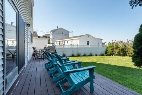 Backyard with wooden deck, teal Adirondack chairs, and a grassy lawn. White houses and a fence are visible in the background under a clear blue sky.