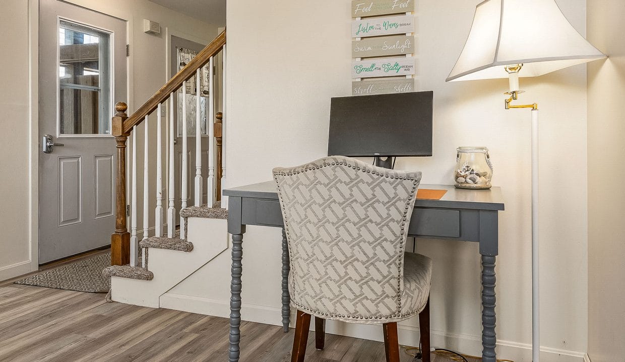 Home office area with a gray desk, computer monitor, and upholstered chair. A nearby lamp provides light. Stairs and a closed entry door are visible in the background.