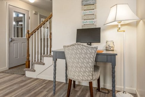 Home office area with a gray desk, computer monitor, and upholstered chair. A nearby lamp provides light. Stairs and a closed entry door are visible in the background.