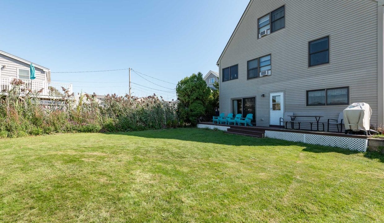 A spacious backyard with a lawn, deck, outdoor seating, and a grill. The house is beige with multiple windows. Some plants and a neighboring house are visible.