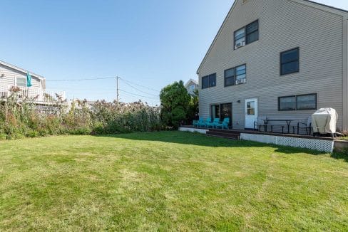 A spacious backyard with a lawn, deck, outdoor seating, and a grill. The house is beige with multiple windows. Some plants and a neighboring house are visible.