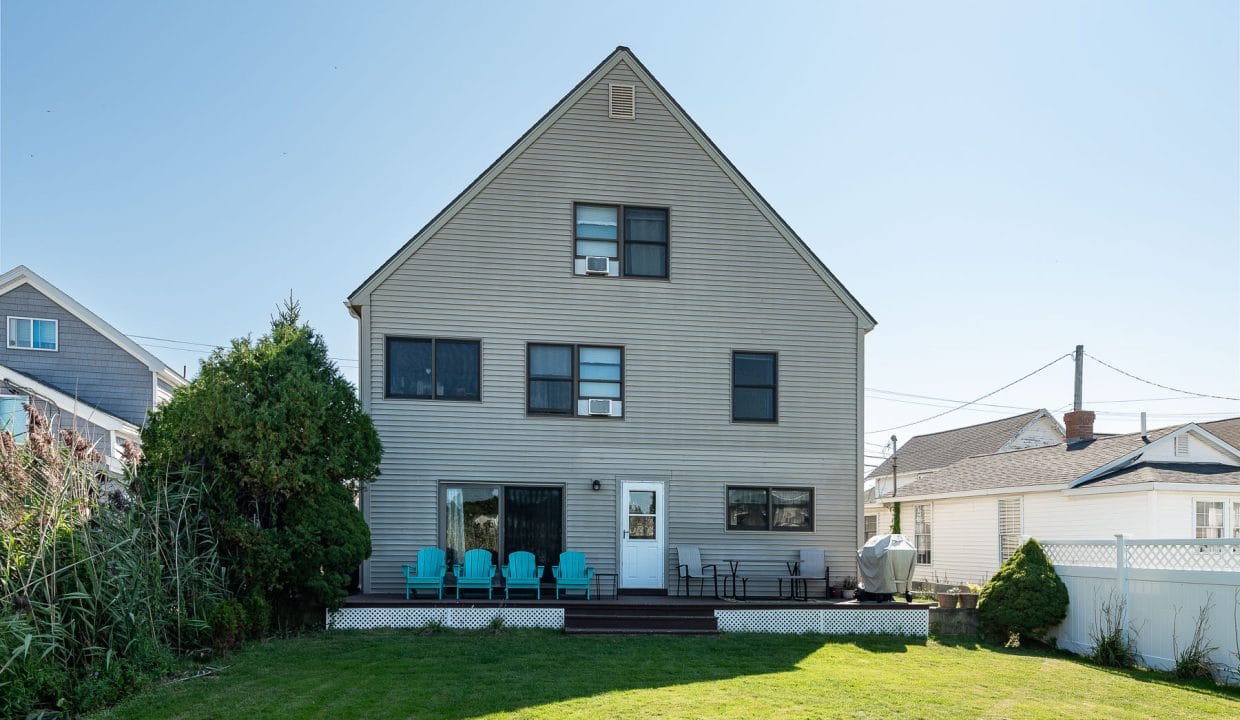 Two-story beige house with a sloped roof, large windows, and a wooden deck with blue chairs. Surrounded by green grass and neighboring houses under a clear blue sky.