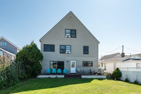 Two-story beige house with a sloped roof, large windows, and a wooden deck with blue chairs. Surrounded by green grass and neighboring houses under a clear blue sky.