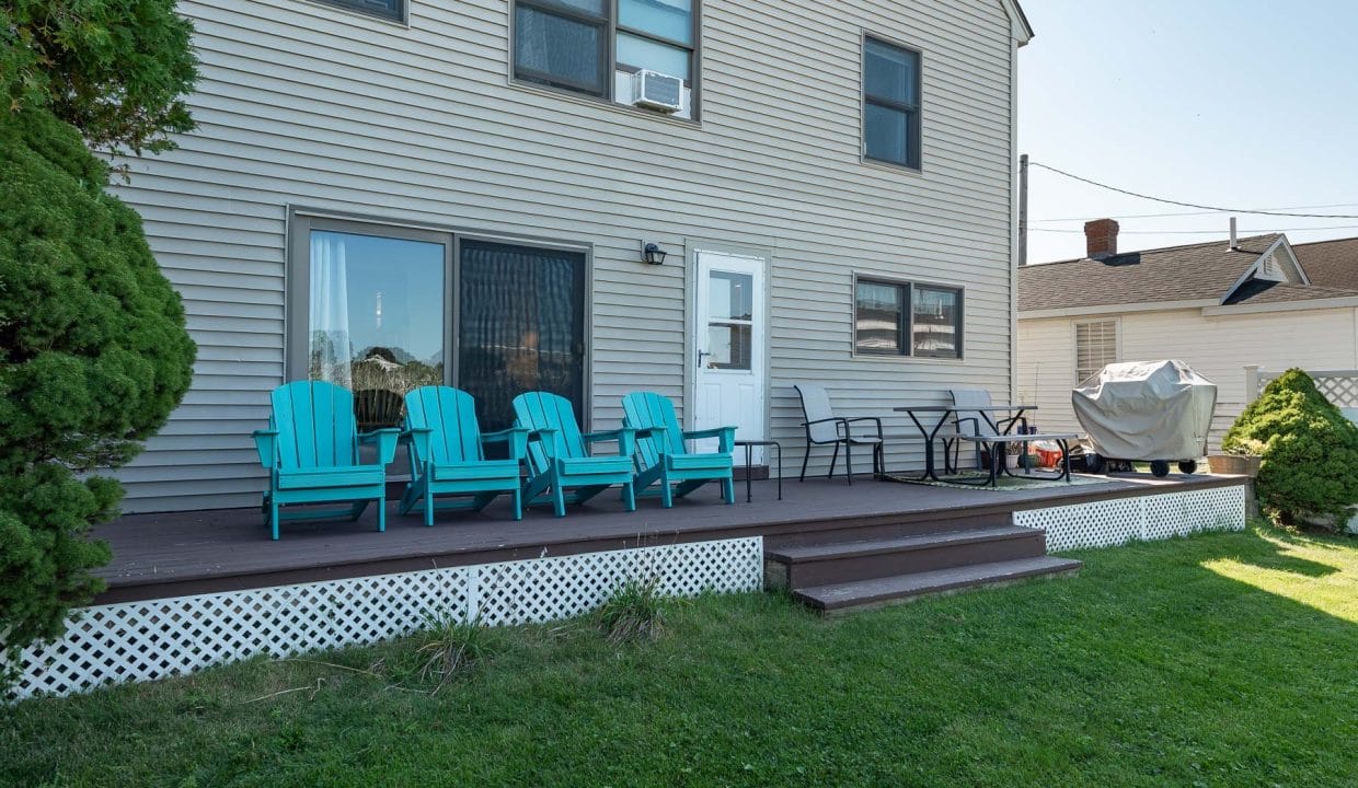 Backyard patio with turquoise Adirondack chairs, a table, and a covered grill. Steps lead to a grassy area.