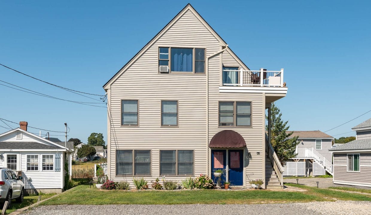 A three-story beige house with a triangular roof, blue door, and small balcony. It has surrounding grass and neighboring houses under a clear blue sky.