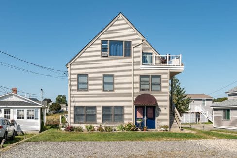 A three-story beige house with a triangular roof, blue door, and small balcony. It has surrounding grass and neighboring houses under a clear blue sky.