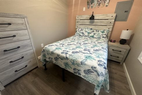 A small bedroom with a wooden bed, matching nightstand, and dresser. The bed is covered with a tropical-themed quilt. Wall decorations include three surfboards and an electrical panel.