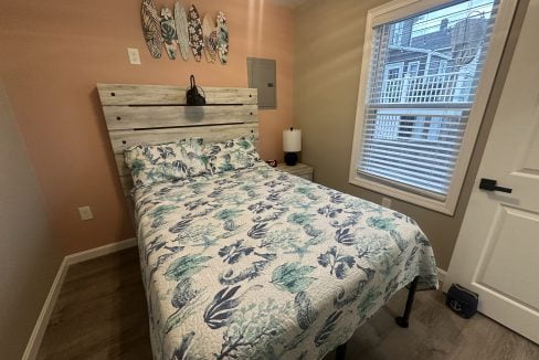 A bedroom with a bed dressed in a blue and green floral comforter, a nightstand with a lamp, wall decor above the headboard, a window with closed blinds, and a door to the right.