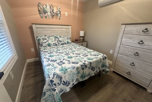 A bedroom with a bed featuring a sea-themed bedspread, a wooden headboard, a bedside table with a lamp, wall-mounted air conditioning, and a wooden dresser. Three fish decorations hang on the wall.