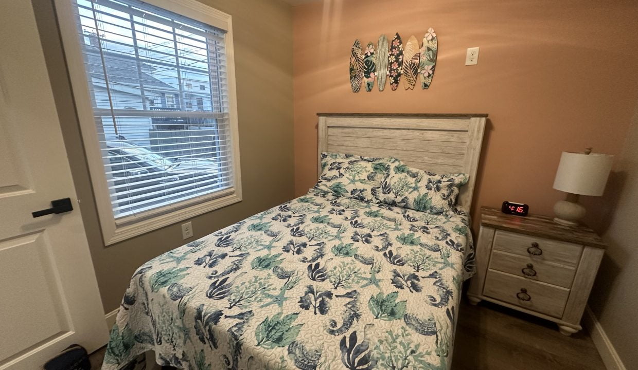 A bedroom with a bed featuring a floral quilt, a nightstand with a lamp and clock, and decorative wooden fish wall art above the bed, near a window with blinds partially open.