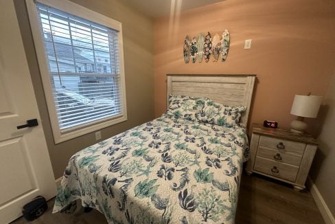 A bedroom with a bed featuring a floral quilt, a nightstand with a lamp and clock, and decorative wooden fish wall art above the bed, near a window with blinds partially open.