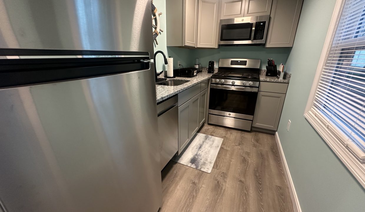 A small, modern kitchen with a stainless steel refrigerator, oven, and microwave, light grey cabinets, granite countertops, a sink, a rug, and a window with blinds.