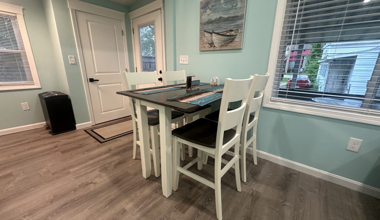 A small dining area with a dark wooden table and white chairs, set against a light blue wall with nautical decor. A window with blinds and a door are visible in the background.