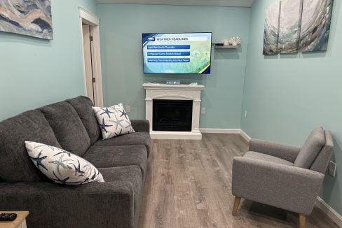 A modern living room with teal walls features a gray sofa, an armchair, nautical-themed pillows, a wall-mounted TV displaying weather headlines, a white fireplace, and coastal decor.