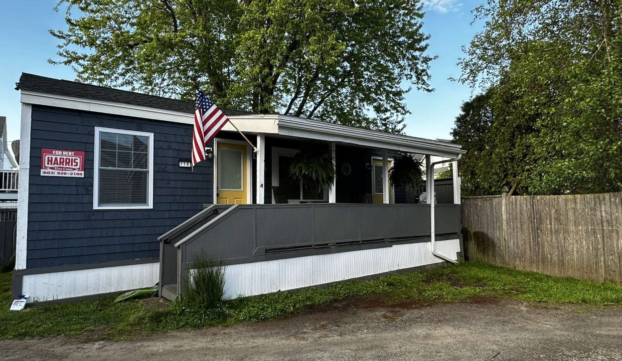 Single-story blue house with an American flag and a for sale sign in the front window. A tree and a wooden fence are in the background. A gravel driveway is in the foreground.