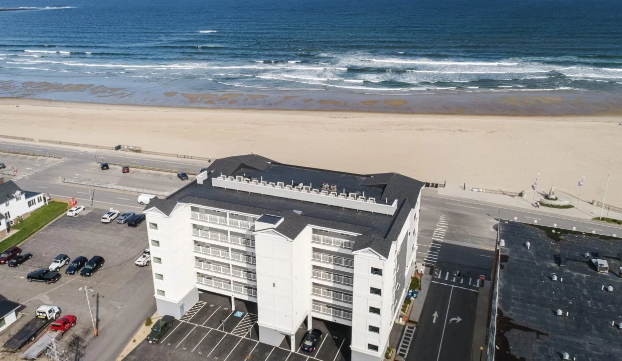 Aerial view of a white multi-story building situated near a beach with a parking lot in front. The ocean and waves are visible in the background, along with scattered parked cars and adjacent buildings.