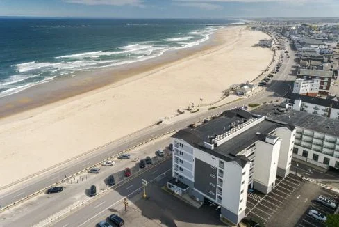 Aerial view of a coastal town featuring a sandy beach, ocean waves, and a road with parked cars parallel to the shoreline. Several buildings are visible along the road.