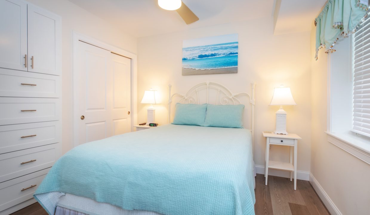 A bedroom with a white bed, light blue bedding, two bedside tables with lamps, a ceiling fan, built-in drawers, and a window with blinds. A beach scene painting is hung above the bed.