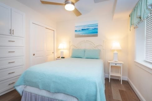 A bedroom with a white bed, light blue bedding, two bedside tables with lamps, a ceiling fan, built-in drawers, and a window with blinds. A beach scene painting is hung above the bed.