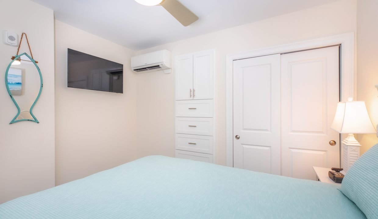 A bedroom with a light blue bedspread, wall-mounted TV, air conditioner, mirror, and a ceiling fan. The room features a white dresser, a closed double-door closet, and a bedside lamp.