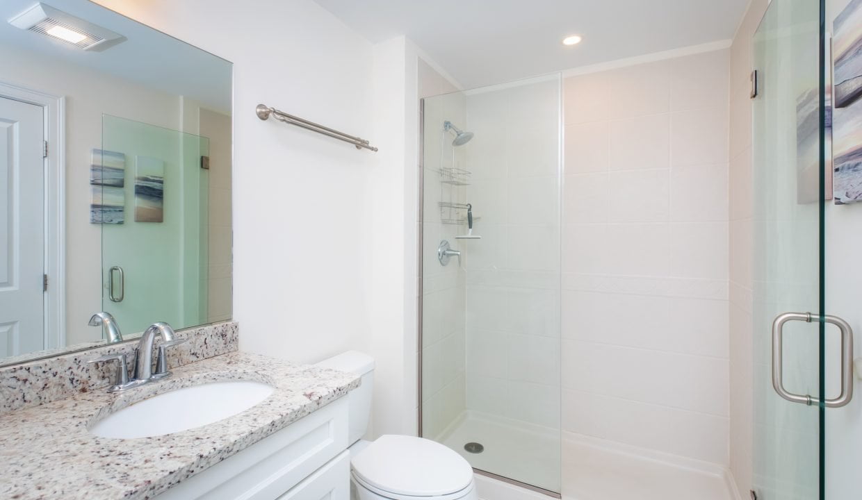 A modern bathroom features a granite countertop with a sink, a toilet, and a glass-enclosed shower. The walls are white, and there is a framed painting next to the shower.