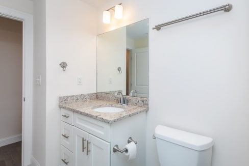 A bathroom with a single-sink vanity, granite countertop, large mirror, overhead lighting, white cabinetry, towel rack, and a toilet with a roll of toilet paper.