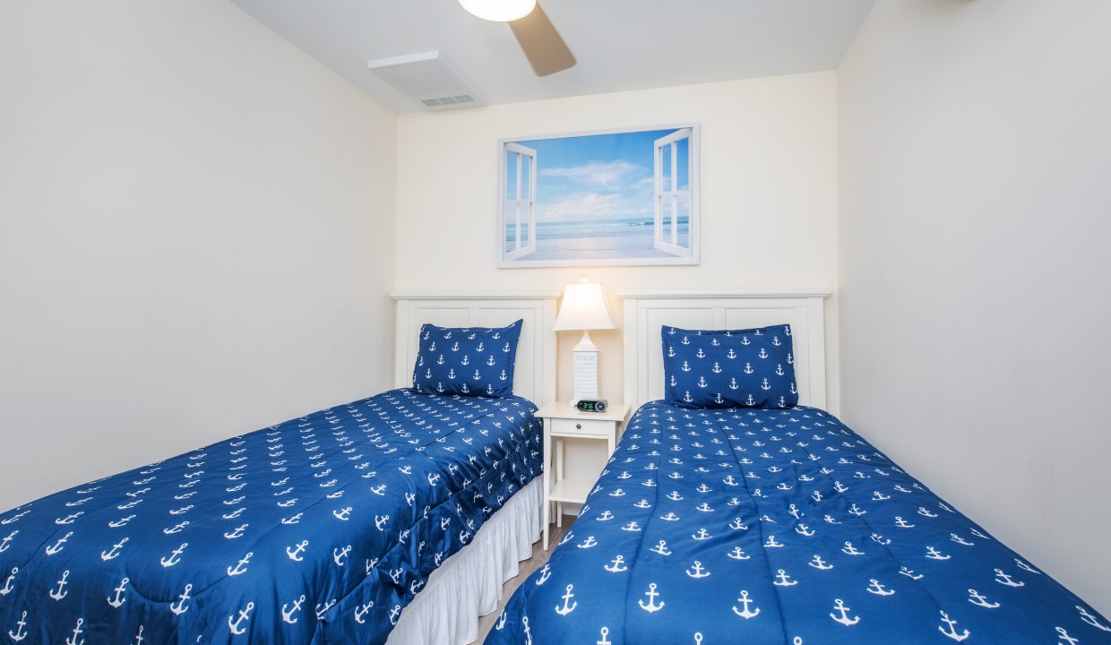 A bedroom with two twin beds covered in blue anchor-themed bedding and a white nightstand with a lamp and clock between them. A ceiling fan is above and a beach-themed picture is on the wall.