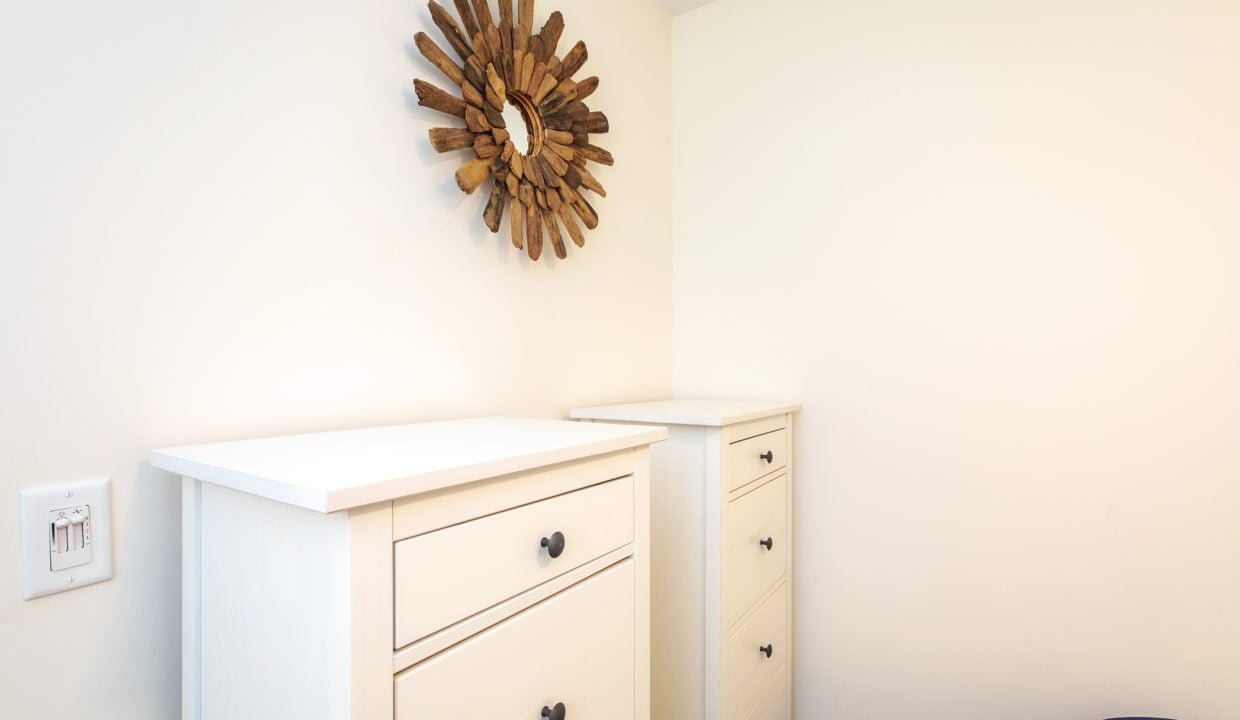 Two white dressers with black knobs are positioned in a corner. A sunburst-shaped wooden wall decoration hangs above, and a light switch is visible on the left.