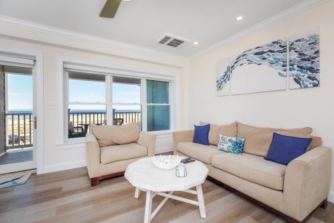 A brightly lit living room with a beige sofa and armchair, blue accent pillows, white coffee table, abstract wall art, and large window showing an ocean view.