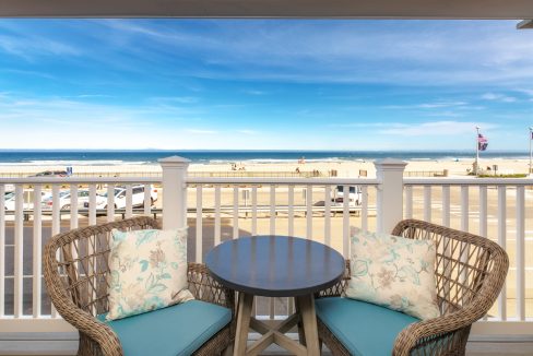 Outdoor patio with wicker chairs, blue cushions, and a round table on a balcony overlooking a sandy beach with calm ocean waves and a clear blue sky. Potted plants add a touch of greenery.