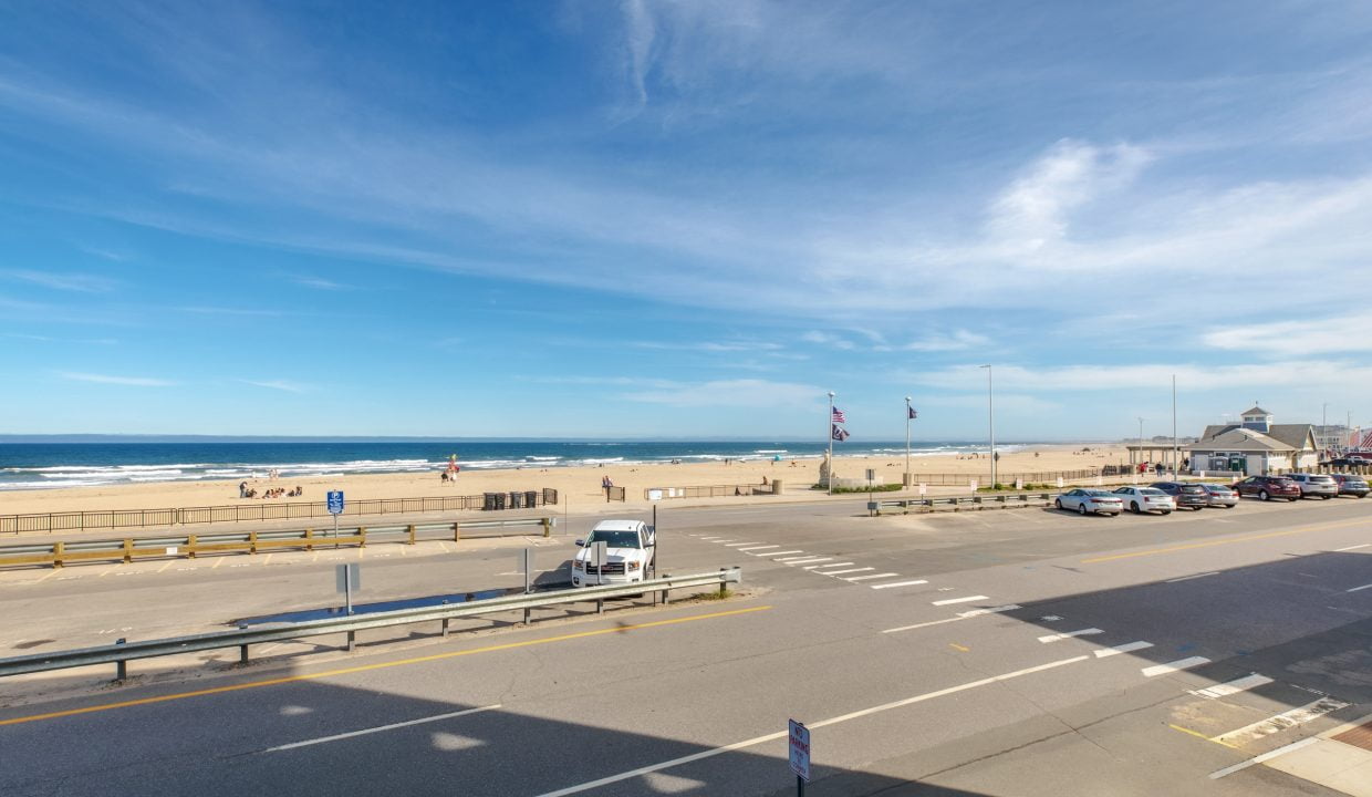 View of a beach with a blue sky and ocean waves, seen from across a road with vehicles and a pedestrian crossing. Flags are flying near a building on the right side.