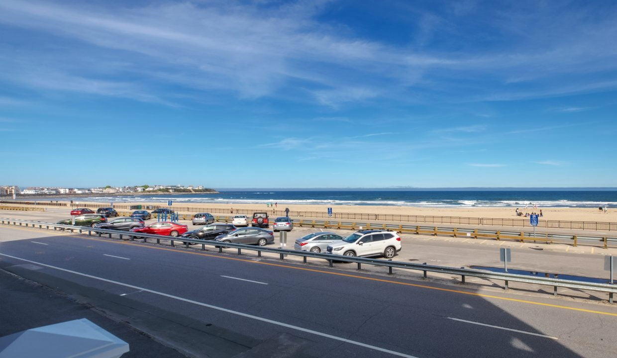 A view of a beach parking lot with several cars parked, a road in front, and the ocean in the background under a clear blue sky. A few people are walking near the shoreline.