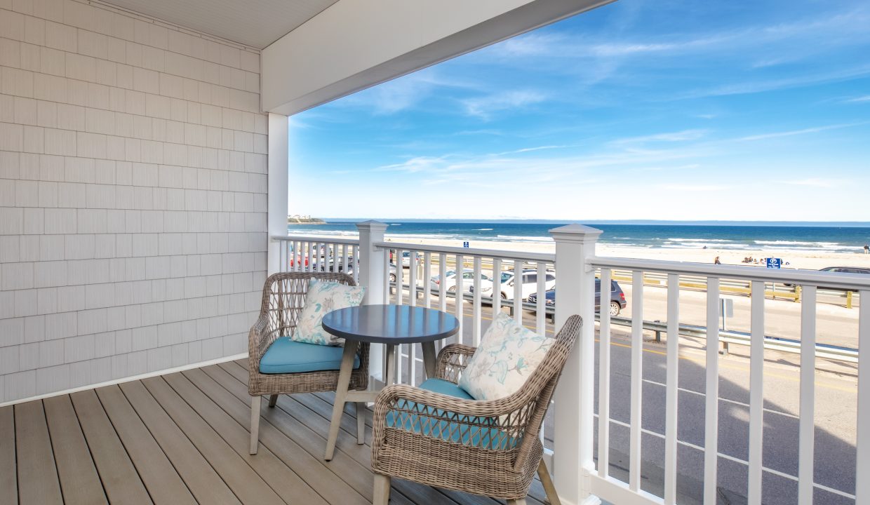 A seaside balcony with two wicker chairs and a small round table overlooks the beach and ocean under a clear blue sky.