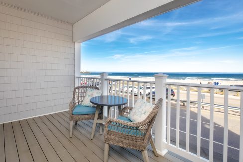 A seaside balcony with two wicker chairs and a small round table overlooks the beach and ocean under a clear blue sky.