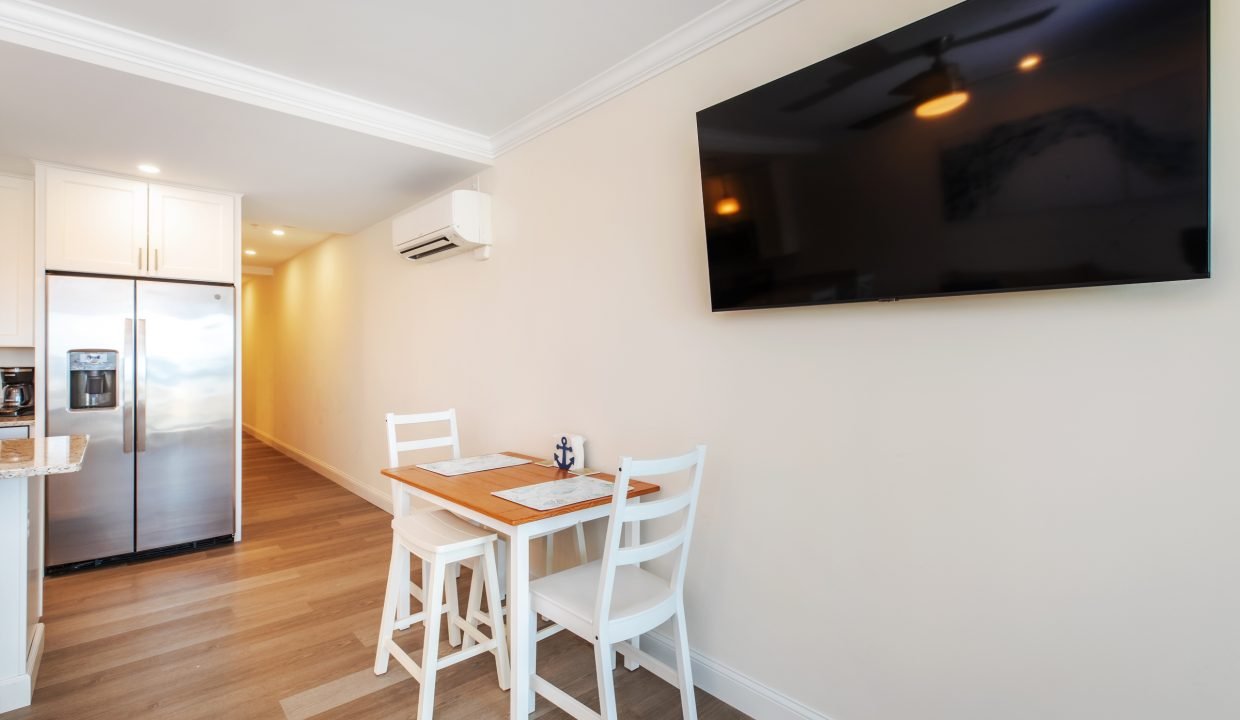 A small dining area with a wooden table and two white chairs is set against a wall. A large flat-screen TV is mounted on the opposite wall. The kitchen has a stainless steel refrigerator.