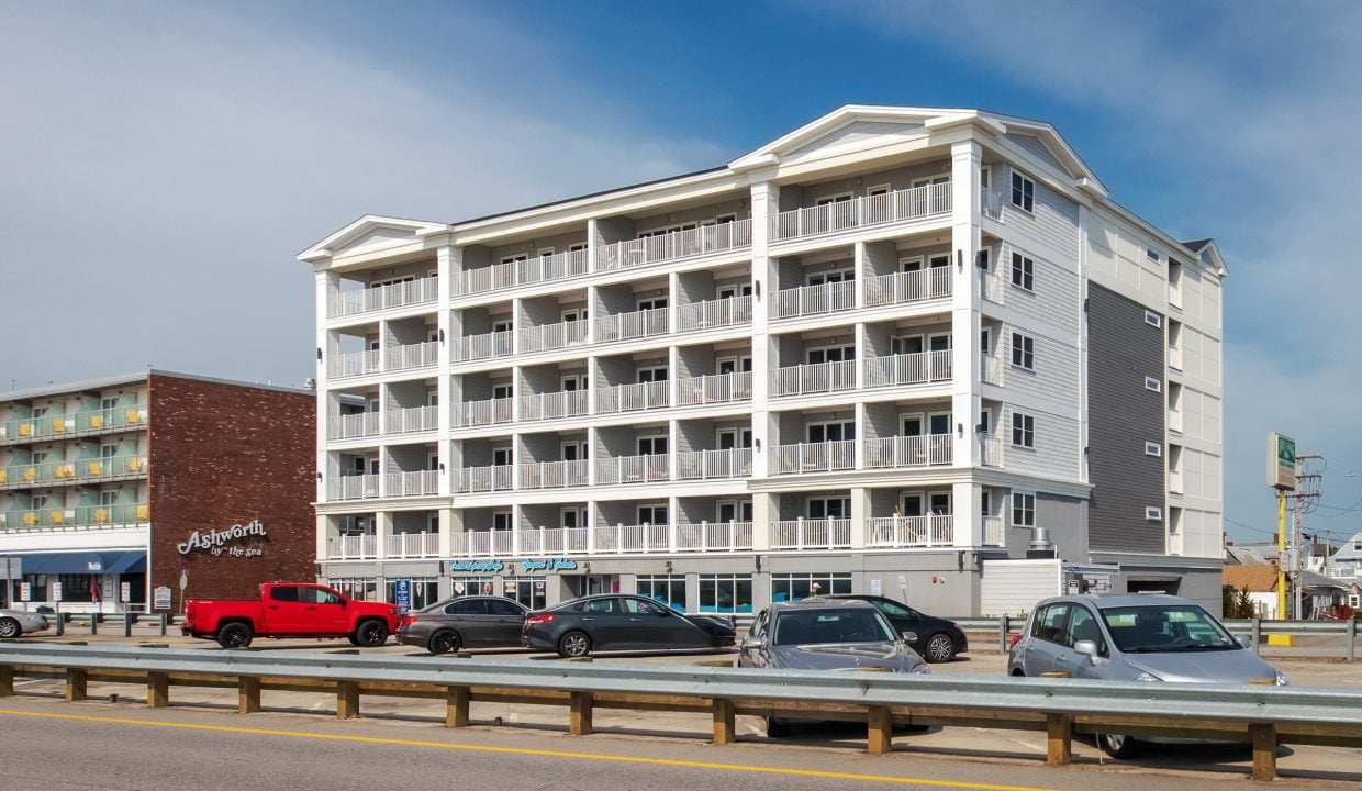 A four-story beachfront hotel with balconies in front of a street with parked cars. A restaurant named 'Angler's' is adjacent on the left. The sky is clear.