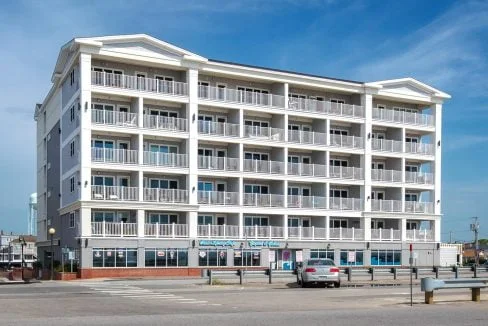 A modern, multi-story beachside building with numerous balconies; storefronts on the ground level; parked cars in front; bright sunny day with a clear blue sky.