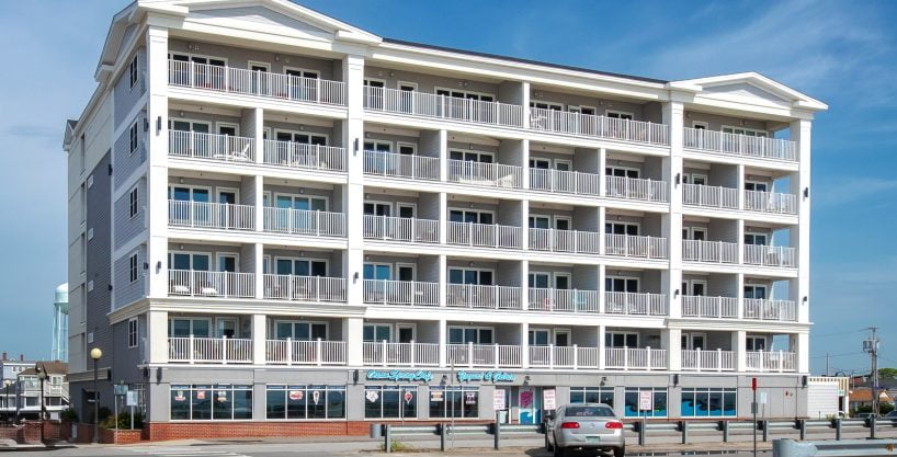A modern, multi-story beachside building with numerous balconies; storefronts on the ground level; parked cars in front; bright sunny day with a clear blue sky.