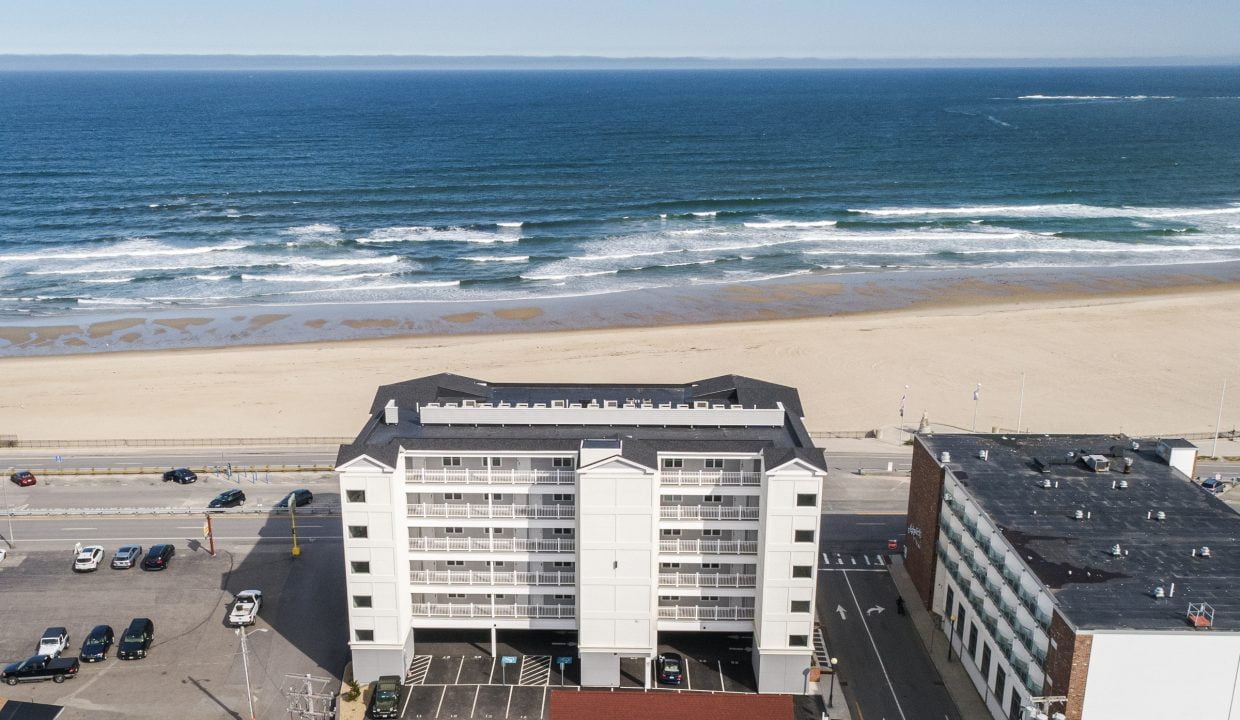 Aerial view of a beachfront building facing the ocean, with a sandy beach and parked cars visible in the foreground. Waves can be seen crashing in the distance.