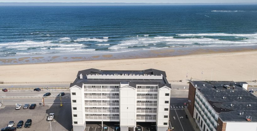 Aerial view of a beachfront building facing the ocean, with a sandy beach and parked cars visible in the foreground. Waves can be seen crashing in the distance.
