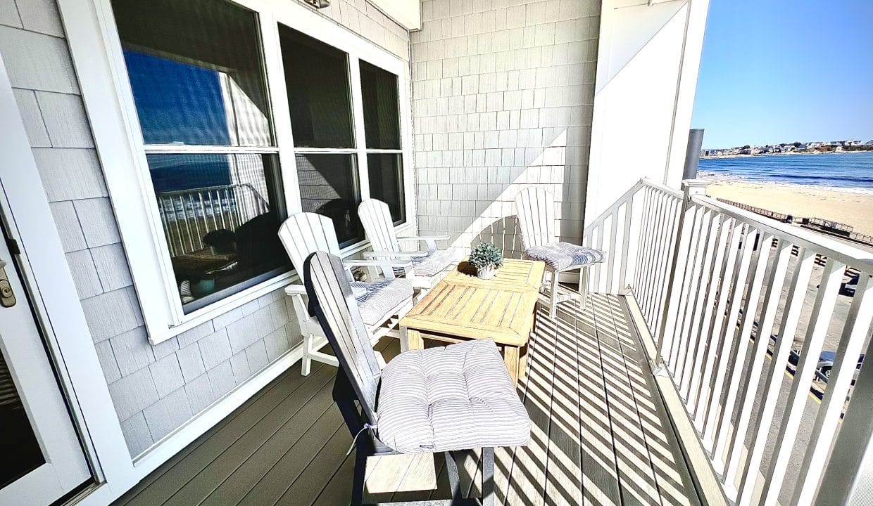 Balcony with white Adirondack chairs and a small table, overlooking a beach and ocean under a clear blue sky.