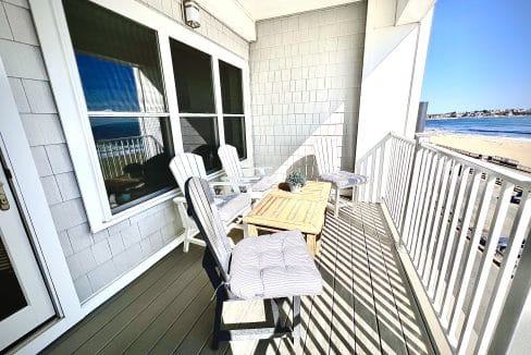 Balcony with white Adirondack chairs and a small table, overlooking a beach and ocean under a clear blue sky.